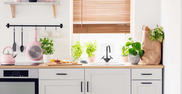 Real photo of a kitchen cupboards, countertop with plants, food, and shopping bag, and window with blinds in a kitchen interior